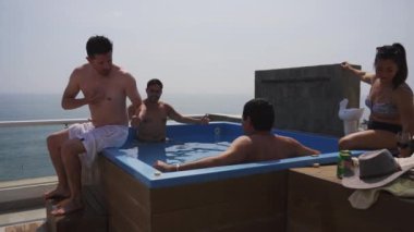 Güneşli bir günde, arka planda deniz olan bir sahil evinin terasında birkaç arkadaşıyla havuzda içki içen bir adam.