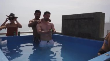 Güneşli bir günde arka planda deniz olan bir sahil evinin terasında havuzda omuz masajı yapan birkaç arkadaş.