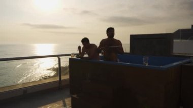 Birkaç arkadaş, güneşli bir günde, güneş batmadan önce deniz manzaralı bir sahil evinin terasında havuzda yemek yiyorlar.