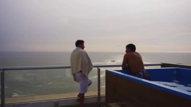 İki arkadaş konuşuyor ve denize bakıyor. Plaj evinin terasında havuzda bira içiyorlar. Bulutlu bir günde.