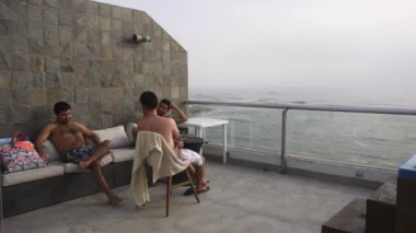 İki arkadaş sahildeki evin terasında oturup konuşuyor ve bira içiyor. Arka planda deniz ve bulutlu bir günde 4K 'da dalgalar var.