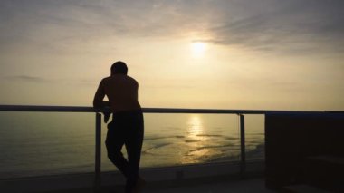 Sahildeki bir evin terasında gün batımını seyreden deniz dalgaları karşısında kameraya şakalar yapan bir adam.