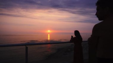 Bir çift güzel, mor bir gün batımına bakıyor. Deniz ve arka plandaki dalgalar güneşin parlaklığını yansıtıyor. 4K 'daki bir sahil evinin terasında.