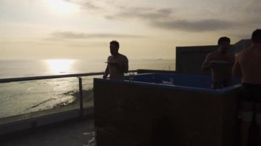 Hızlı kamera arkadaşları, arka planda güneş ve denizin parlaklığını yansıtan dalgalarıyla bir sahil evinin terasında içki yiyip içiyorlar. 4k içinde zaman aşımı