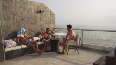 Birkaç arkadaşın terasta oturup deniz arka planı ve gün içinde dalgalarla dolu bir sahil evinde bira içerken çekilmiş bir fotoğraf makinesi. 4k içinde zaman aşımı