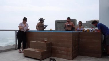 Birkaç erkek ve bayan arkadaşın terasta bir havuzda bira yiyip içerken ve deniz dalgalarının arka planında bir sahil evinde sohbet ederken çekilmiş bir kamera. 4k içinde zaman aşımı