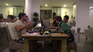 Birkaç arkadaş ve kız arkadaşları, dizüstü bilgisayarlarıyla oynamadan önce oturma odasında bira içip kadeh kaldırıyorlar.