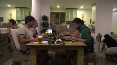 Birkaç arkadaş yemek masasında dizüstü bilgisayarlarıyla oynuyor, bira içiyor ve kız arkadaşları ve kız arkadaşları oturma odasında yerde oyun oynuyorlar.