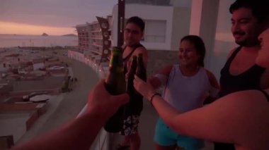 POV 'un ilk bakışı, erkek ve kadın arkadaşlarıyla kadeh kaldırırken elinde bir şişe bira ve deniz üzerinde güneş görünen bir sahil evinin terasından güzel bir gün batımı olan bir şişe bira.