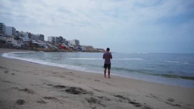 Arkada duran bir adam deniz kıyısında kumlu, dalgalı ve binalı cep telefonuna bakıyor. Soyutlama, barış, rahatlama, toplama kavramı