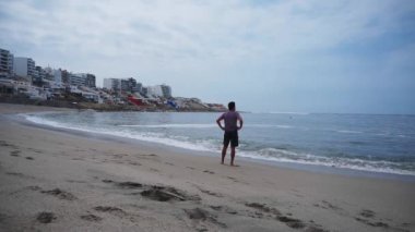 Arkadan bir adam deniz kıyısında yüzmeye ve koşmaya hazırlanan kum, dalga ve binalarla polo tişörtünü çıkarıyor. Soyutlama, barış, rahatlama kavramı