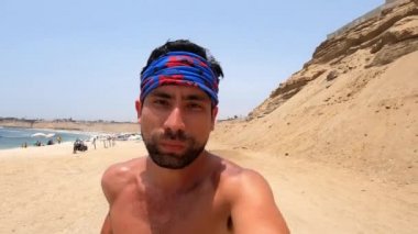 Ciddi bir adamın özçekim portresi. Sahilde deniz kenarında koşan kameraya bakıyor ve insanlar gün içinde güneşleniyorlar.