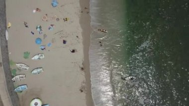 Sahilde şemsiyelerle, oyun oynayan, kristal berrak suda kumsalda kayalarla yıkanan insanların yukarıdan insansız hava aracı görüntüsü 4K 'da kuş bakışı dalgaların yanında.