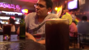 Bir bar masasında, arka planda ortağıyla sohbet eden ve 4K 'de bira içen bir adamla Küba libresinin yakın çekimi.