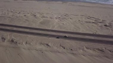 Çölün ortasında bir arabayı takip eden hava aracının hava görüntüsü ve günün şiddetli rüzgarı nedeniyle hareket eden kumla birlikte deniz 4K 'lık yoğun güneşle birlikte.