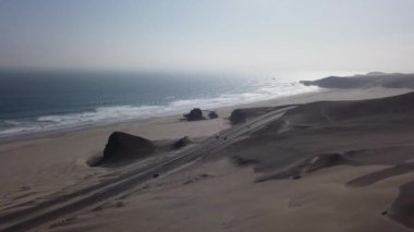 Çölde arabalı bir otobanın hava aracı görüntüsü kum tepeleri ve dalgalı deniz ve bulutlu bir günde sahilde kum.