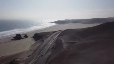 Çölde arabalı bir otobanın hava aracı görüntüsü kum tepeleri ve dalgalı deniz ve bulutlu bir günde sahilde kum.