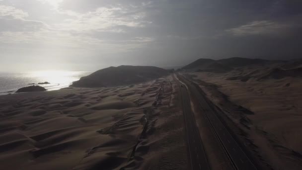 海と道路の間に砂がある美しい色の原生砂漠に降下する空中ドローンの眺め4Kの日に山と砂丘がある車 — ストック動画