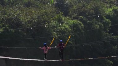 Tanımlanamayan bir çift, renkli basamakları ve pek çok ağacın arka planında asma köprüde yürüyor.