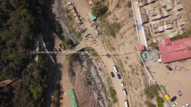 Bir asma köprüden aşağıya bakan insansız hava aracı dağların yamaçlarında farklı renkli bir nehirde yürüyen insanlar ve kuş bakışı yeşil ağaçlar.