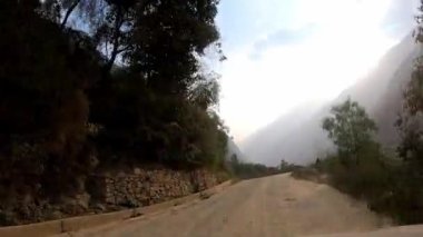 Arabadaki POV, dağlık arazilerdeki dar bir toprak yolda çalılar ve kayaların arasından geçiyor. Dağlar ve yeşil ağaçlar, temiz gökyüzünde güneş görüyor.