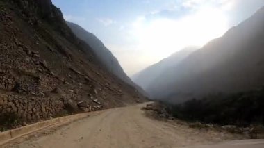 Arabadaki POV hızlı kamerası dağlık arazideki dar toprak bir yolda çalıların ve kayaların arasından geçiyor. Tarlaları, dağları ve yeşil ağaçları var. Güneş açık gökyüzünde.