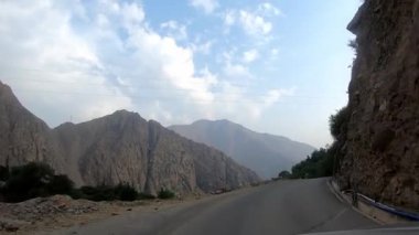 POV dar asfalt yolda, dağların yanı sıra uçurumları olan çalılar ve temiz gökyüzünde güneşli yeşil ağaçlar arasında araba sürüyor.