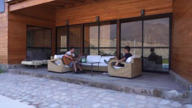 Beyaz bir adam terasta dizüstü bilgisayarı ve gitarıyla sanal müzik dersleri veriyor. Dağlar arasında bir tatil kulübesinde.