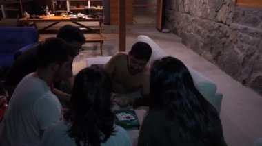 Birkaç arkadaş kadeh kaldırıyor ve kağıt oynuyor. Oyun salonda gergindi.