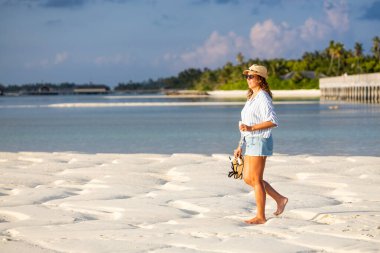 Orta yaşlı bir kadın sahilde bir bardak şarapla yürüyor.