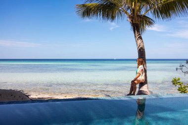 Rahat bir kadın yüzme havuzu ve okyanusun yanındaki bir palmiye ağacına yaslanıyor.