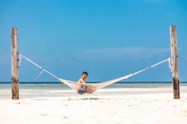 Küçük bir çocuk beyaz bir kumsalda hamakta dinleniyor.