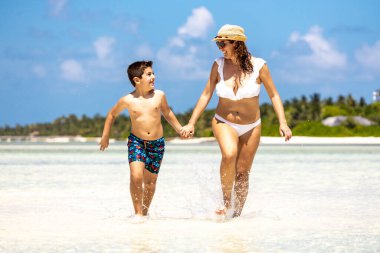 Anne ve oğlu beyaz bir kumsalda suyun içinde koşuyorlar.