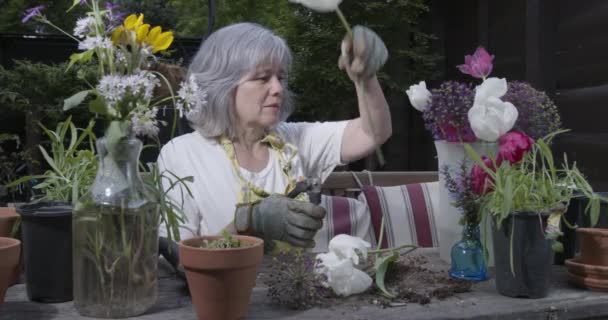 Slider shot of beautiful caucasian senior woman arranging flowers in vase behind wood potting bench outdoors in patio — Stok Video