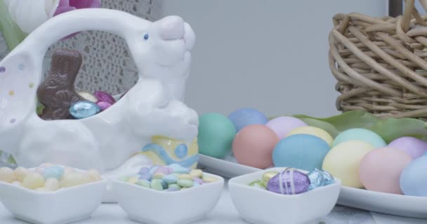 Панельний знімок кольорових великодніх яєць, цукерки в посуді, тюльпани в плетеному кошику, без білої ковдри гачком на фоні — стокове відео