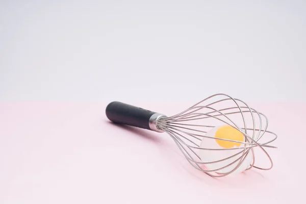Whisk with transparent egg on pastel pink. Light banner background, copy space for text. Culinary mixer, whipping egg white and yolk, preparing and mixing cream.