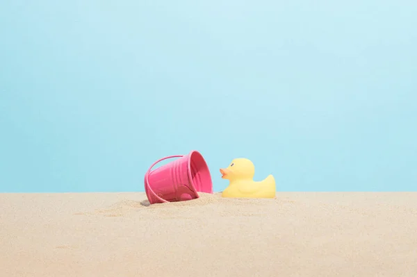 Yellow rubber ducky and pink bucket on the beach sand. Minimal summer vacation composition.