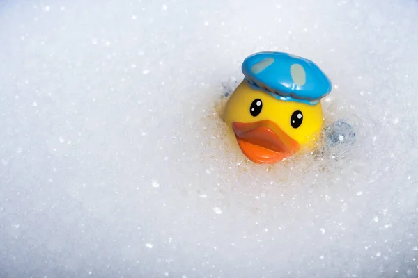 A yellow rubber duck for a bath in a bath cap and with a shower head in the wing in a cloud of soapy foam