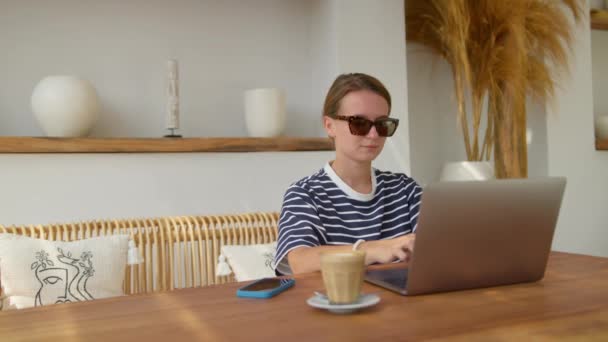 一位面带微笑的年轻女子戴着眼镜 一边与手提电脑一起工作 一边坐在户外餐桌边喝咖啡 — 图库视频影像