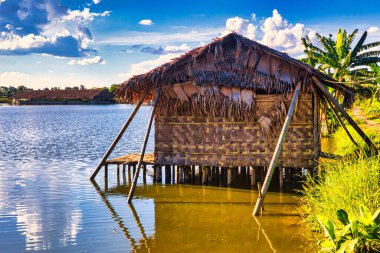 Bago, Myanmar - 02 Aralık 2012: Geleneksel Myanmar bambu kırsal evi ya da kırsal kesimde nehir ve uzun kuyruklu teknelerin bulunduğu cambaz ayaklıkları üzerindeki kulübe