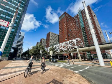 Den Haag, Hollanda - 25 Ağustos 2021: Arka planda modern toplu taşıma araçları ve gökdelenlerle bisiklet yolundan geçen iki bisikletçi