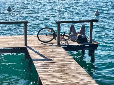 Menthon Saint Bernard, Fransa - 10 Ekim 2021: Bisikletli genç bir adam güneşleniyor ve dağ gölündeki ahşap bir iskelede mola veriyor.