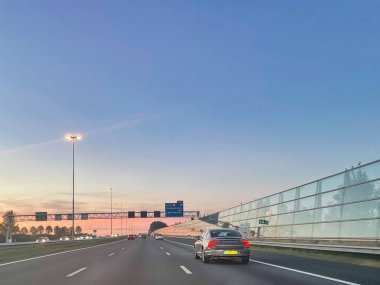 Zevenbergschenhoek, Hollanda - 24 Ekim 2021: arabalar günbatımında modern şeffaf gürültü bariyerleriyle bir otoyolun asfalt şeritlerinde sürüyorlar