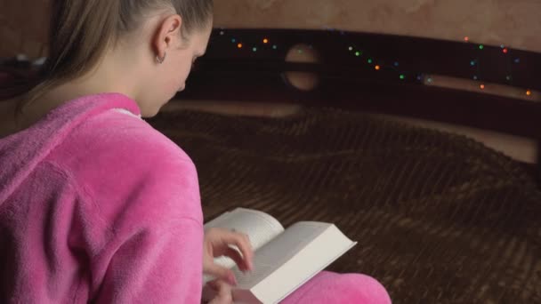 Jonge vrouw in roze pyjama zit leest een boek — Stockvideo