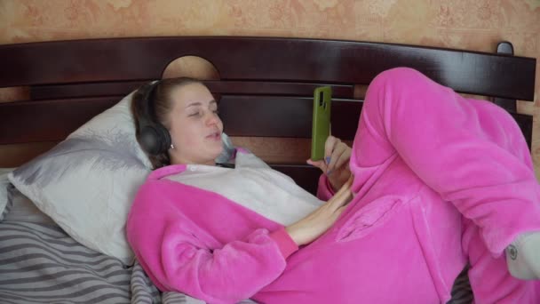Девушка в пижаме на кровати в наушниках разговаривает по видеосвязи по телефону — стоковое видео