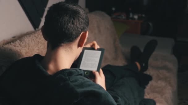 Человек в халате читает электронную книгу, сидя на диване — стоковое видео