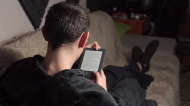 El hombre en un albornoz lee un libro electrónico mientras está sentado en el sofá — Vídeos de Stock