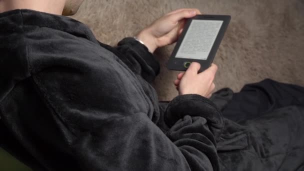 Homem de roupão lê um livro eletrônico enquanto está sentado no sofá — Vídeo de Stock