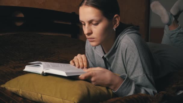 Aranyos lány olvas és leveleket keresztül egy könyvet, miközben fekszik kényelmesen az ágyban — Stock videók
