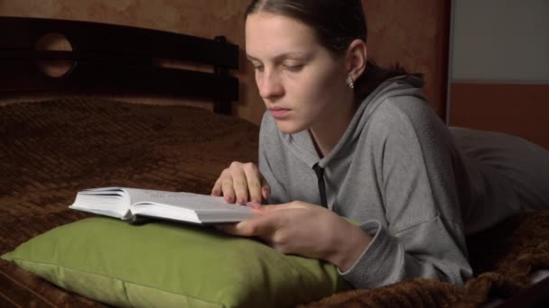 Hermosa chica lee un libro cómodamente mientras está acostado en la cama — Vídeos de Stock